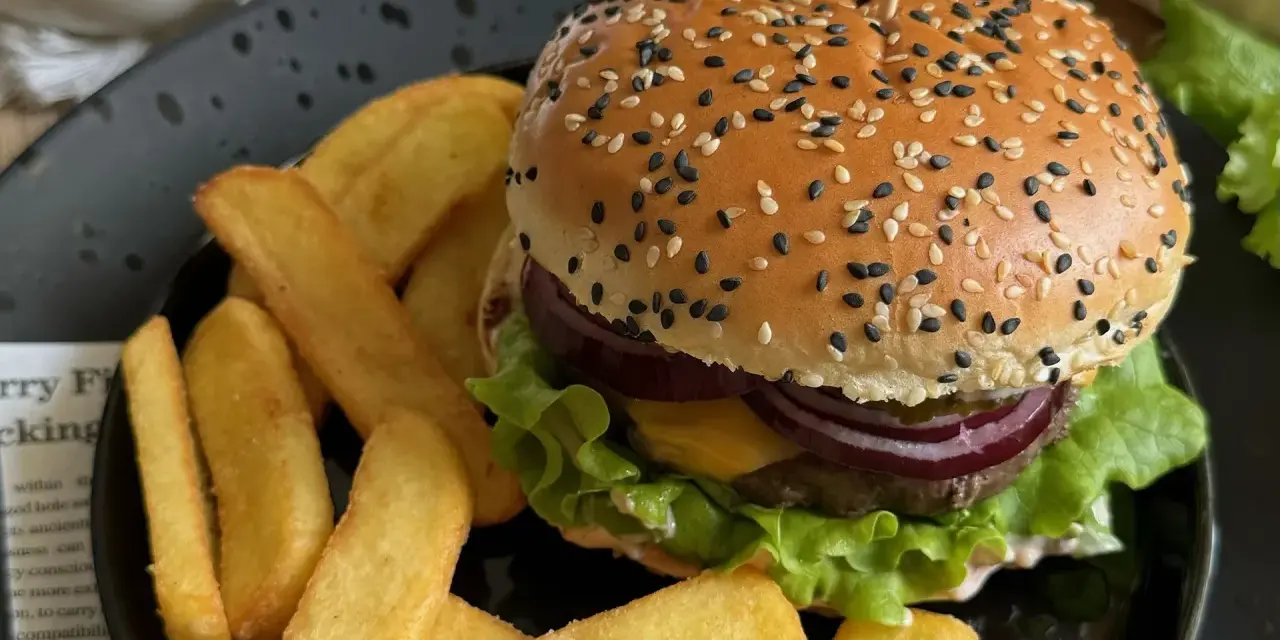 Soczysty burger z sezamem i frytki na czarnym talerzu.