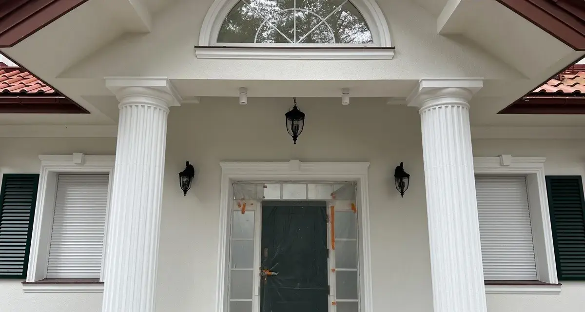Elegancki pilaster zdobi fasadę domu, wspierając dach nad wejściem. Obok okna i drzwi, tworzy klasyczną kompozycję.