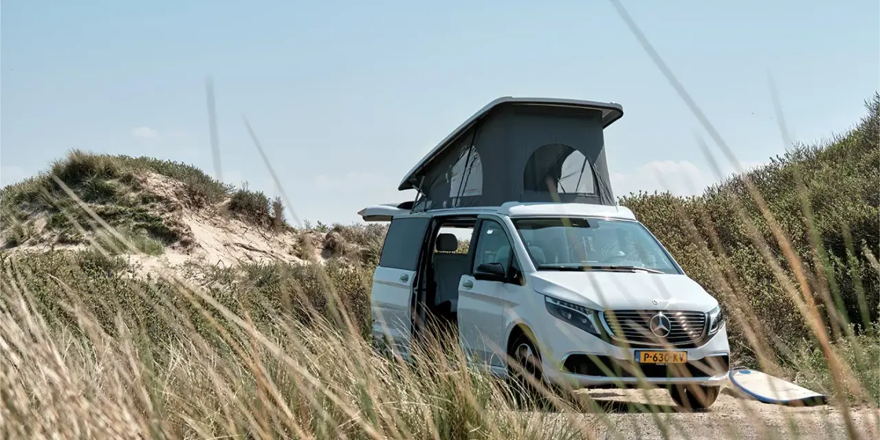Vit el campervan med uppfällt tak står parkerad vid en sanddyn. En surfbräda ligger bredvid.