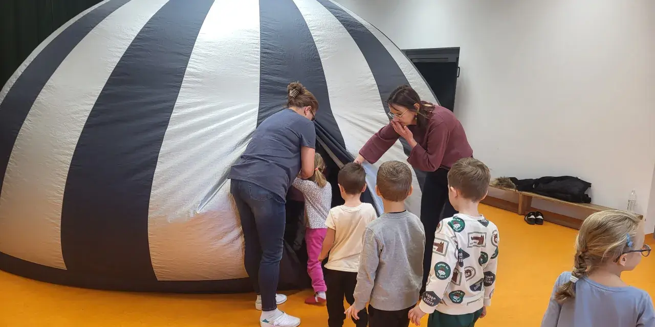 Dzieci i opiekunki wchodzą do dmuchanej kopuły, która przypomina planetarium. To miejsce, gdzie można oglądać filmy z przesłaniem, odkrywając tajemnice wszechświata.