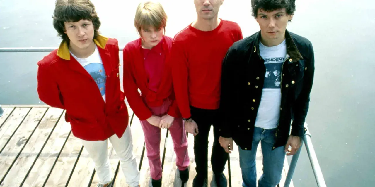 Die Band Talking Heads steht auf einem Holzsteg. David Byrne trägt ein rotes Hemd, Tina Weymouth ein pinkes Outfit.