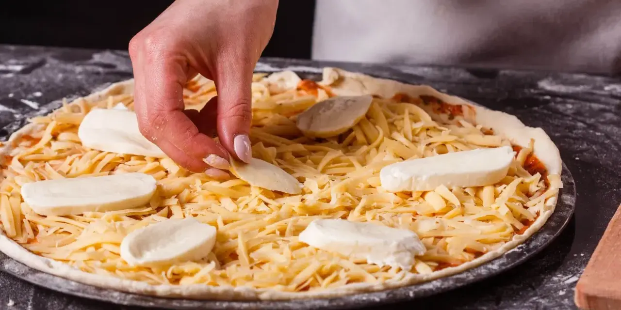 Dłoń układa kawałki sera na pizzy, która jest już posypana startym żółtym serem.