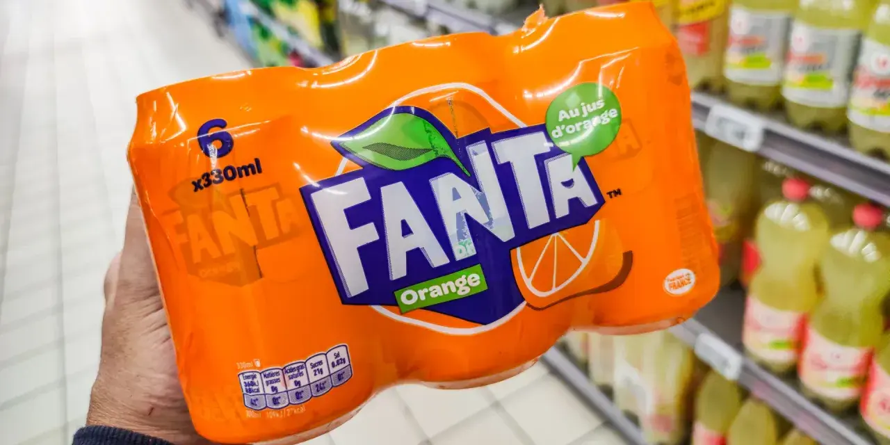 Opakowanie 6 puszek Fanty Orange w ręce, na tle sklepowych półek z napojami.