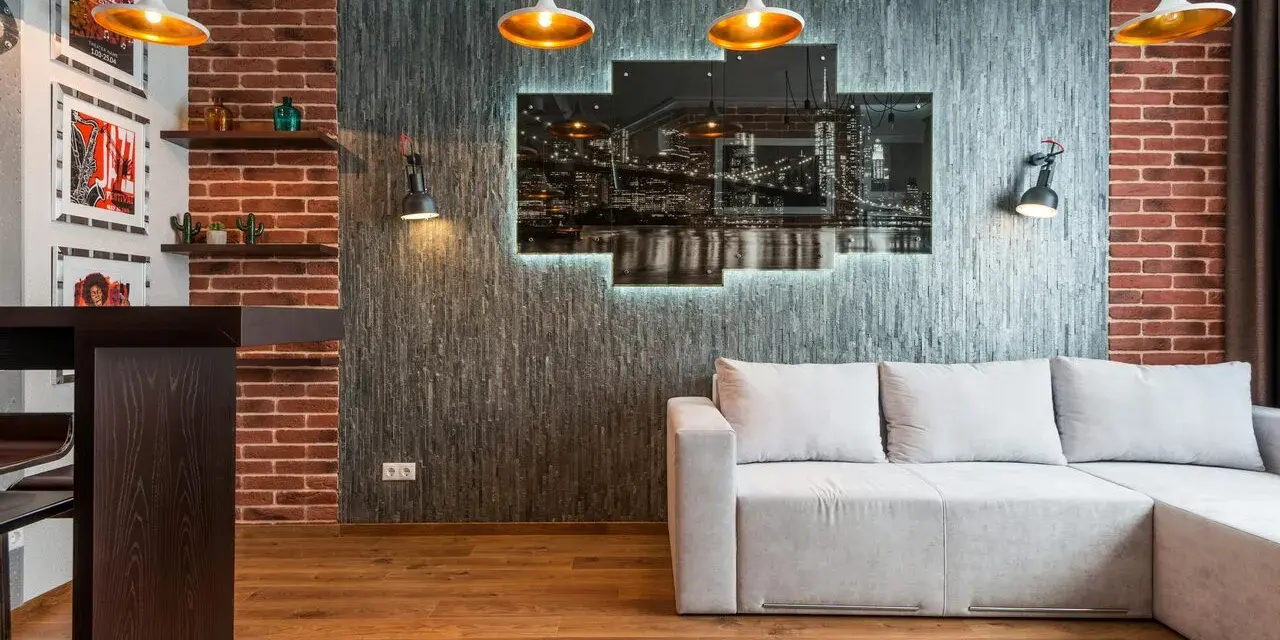 Nowoczesny salon w stylu loft: szara kanapa, ceglana ściana, panel z nocnym widokiem miasta i złote lampy.