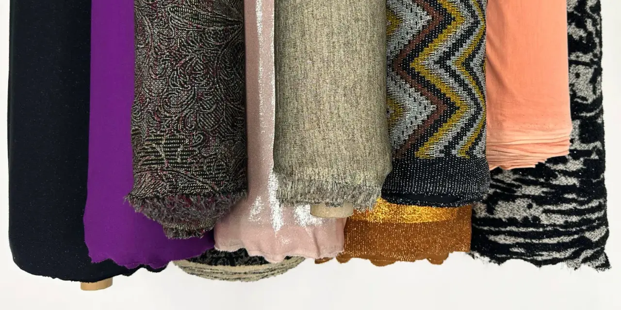 Rouleaux de tissus aux textures variées : noir uni, violet, motifs complexes, rose pâle, beige chiné, zigzags multicolores, pêche, et noir et blanc. Une matière mesh se distingue par sa finesse.