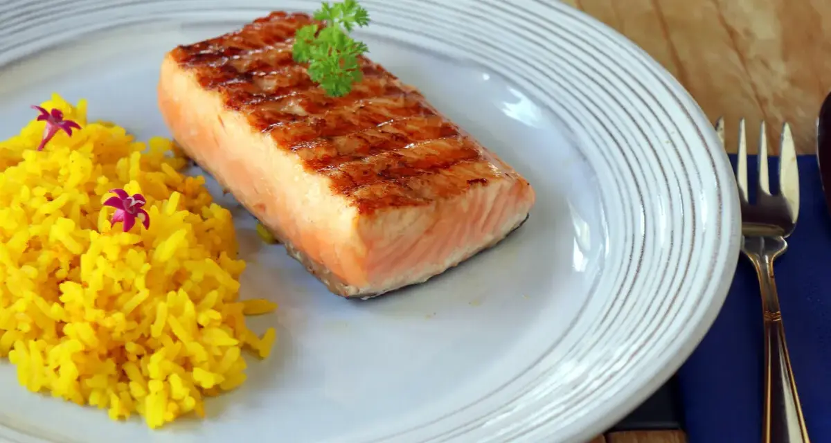 Un delicioso salmón a la plancha con marcas de parrilla, servido con arroz amarillo y decorado con perejil y flores moradas.