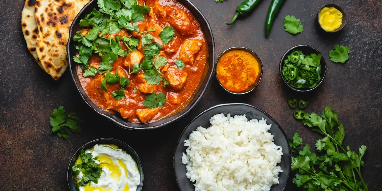 Curry z kurczakiem, ryż, chlebki naan, jogurt i przyprawy na ciemnym tle.