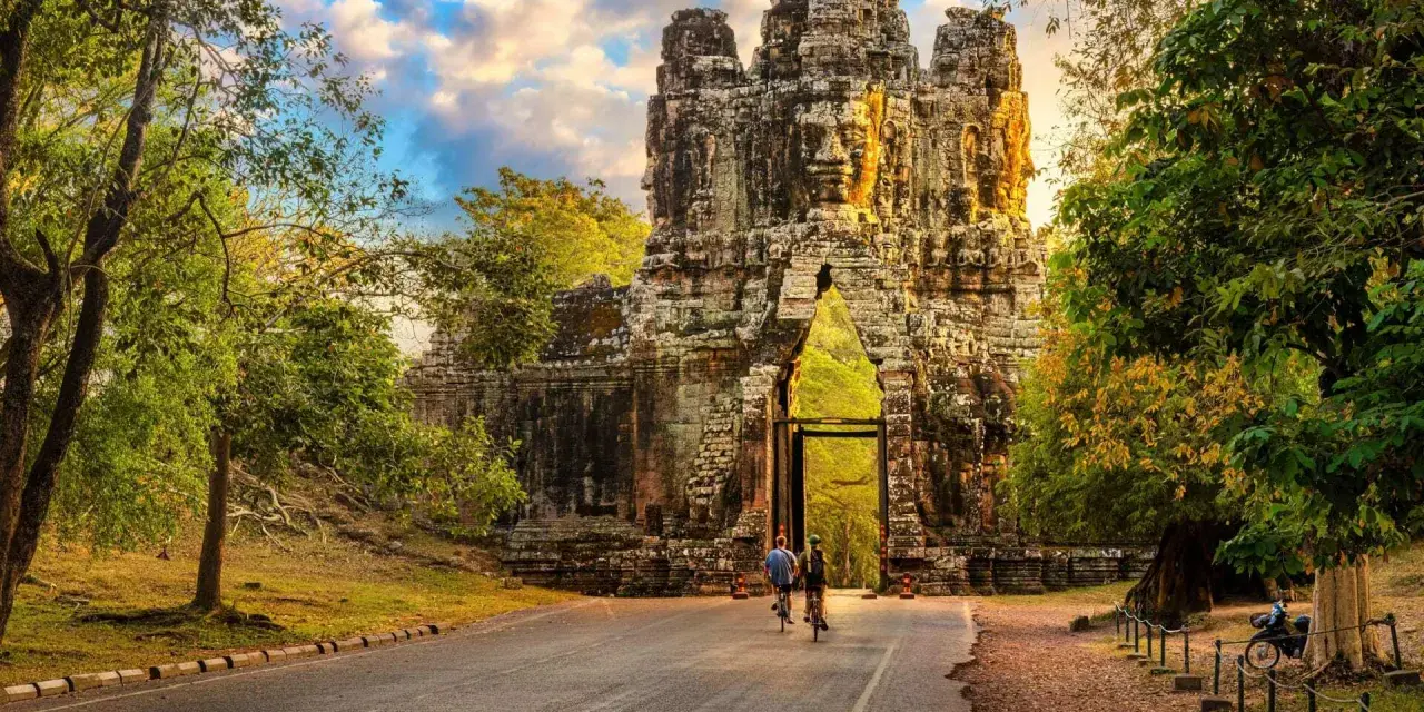 Two cyclists approach a massive stone gateway, one of the ancient Cambodia temples, bathed in golden sunlight.
