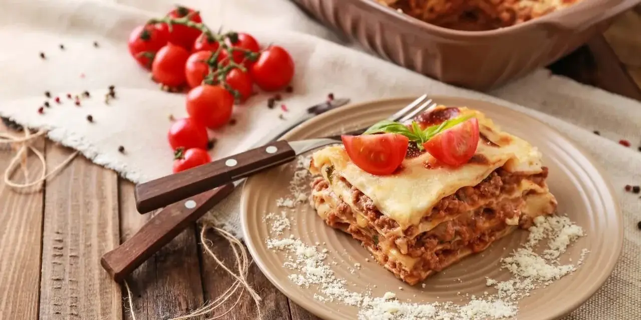 Pyszna, wielowarstwowa lasagne z mięsem mielonym, polana sosem beszamelowym i posypana parmezanem. Na talerzu ozdobiona pomidorkami koktajlowymi i bazylią.