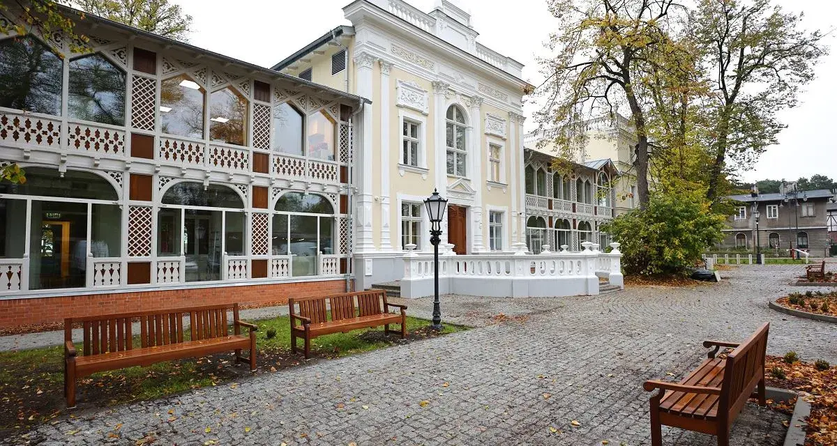 Elegancki budynek z ażurowymi werandami, przypominający dawny **Hevelianum dom zdrojowy**. Brukowany dziedziniec z ławkami i jesiennymi liśćmi.