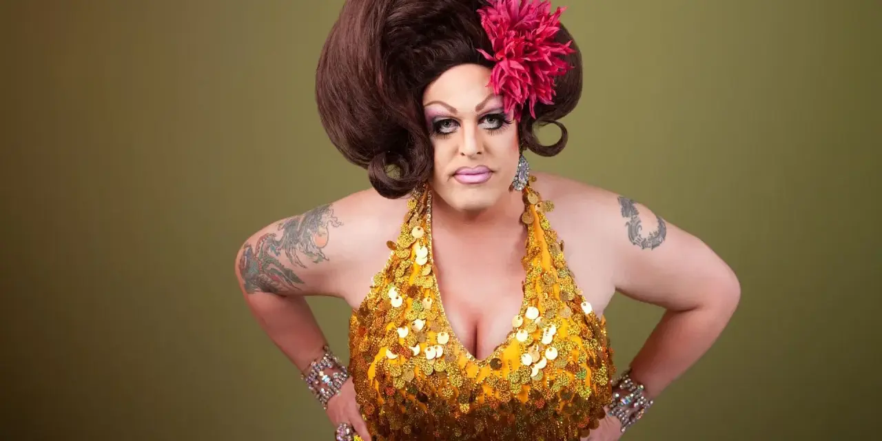 Osoba w złotej, cekinowej sukience i peruce, z kwiatem we włosach. To przykład tego, co to jest drag queen – artystyczna forma ekspresji.