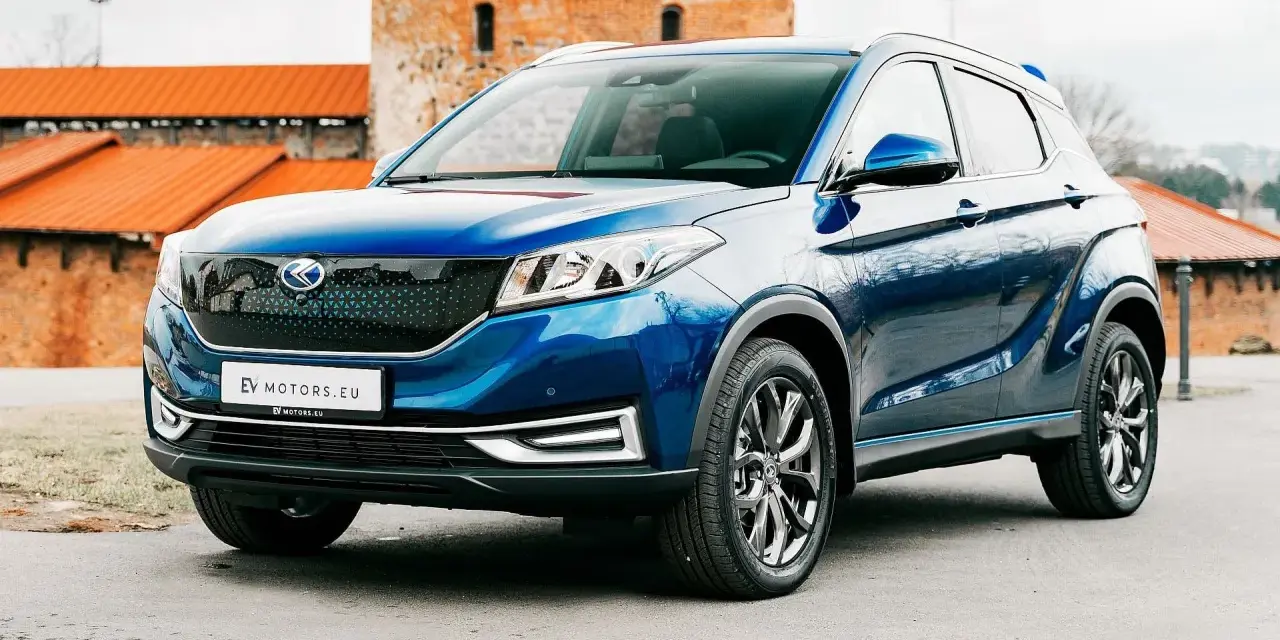 Błękitny SUV EV Motors.eu z charakterystycznym grillem, zaparkowany przed zabytkowym budynkiem z cegły.