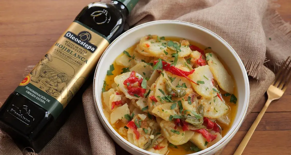 Un tazón de patatas a lo pobre, con pimientos rojos y verdes, perejil picado y una botella de aceite de oliva virgen extra.