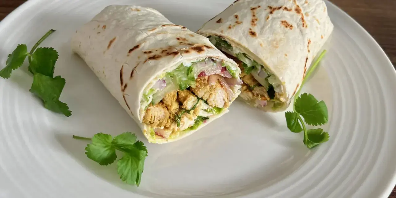 Kebab w tortilli przekrojony na pół, z kurczakiem, warzywami i kolendrą na białym talerzu.