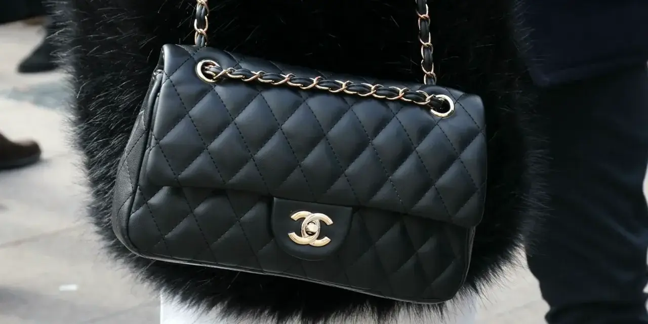 Elegancka, pikowana torebka chanel czarna z złotym łańcuszkiem, noszona na czarnym futrze.