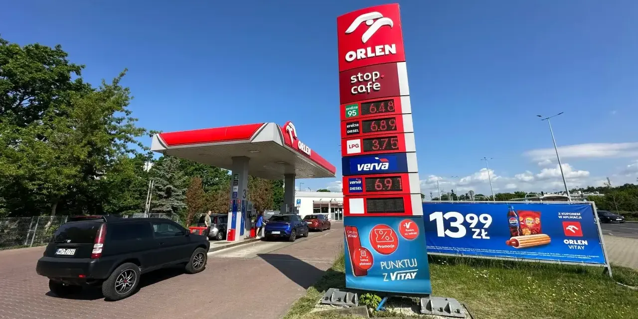 Stacja paliw Orlen z cennikiem paliw: benzyna 95 za 6.48 zł, diesel za 6.89 zł, LPG za 3.75 zł. Obok reklama hot doga i napoju.