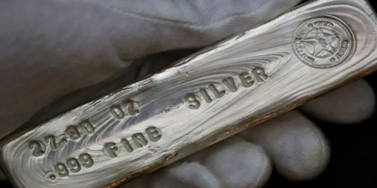 Mano guantata tiene un lingotto di **titolo argento** 999.9, con incisioni che indicano 27.00 oz e "FINE SILVER".