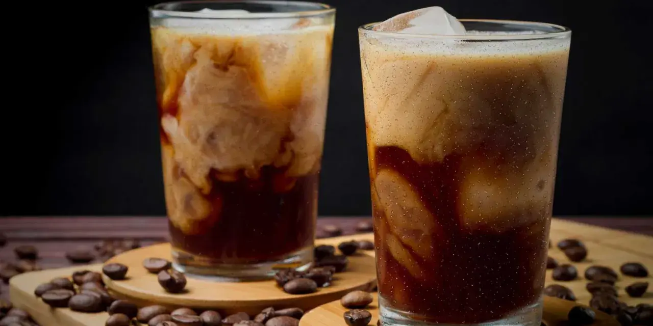 Två glas med iskaffe, där grädden virvlar i det mörka kaffet. Kaffebönor sprider sig runt glasen.