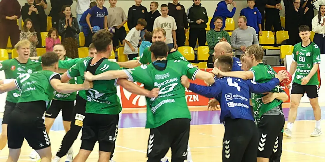 Krystian Woźniak i jego drużyna świętują zwycięstwo. Zawodnicy w zielonych strojach przytulają się na boisku, a na trybunach widać kibiców.