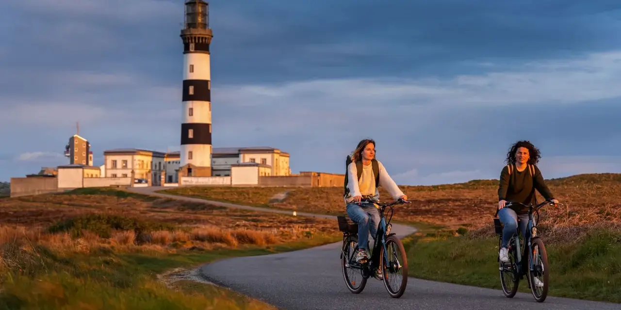Deux femmes font du vélo sur une route sinueuse à Ouessant, avec un phare emblématique en arrière-plan. Une belle façon de visiter Ouessant à pied, ou plutôt à vélo !