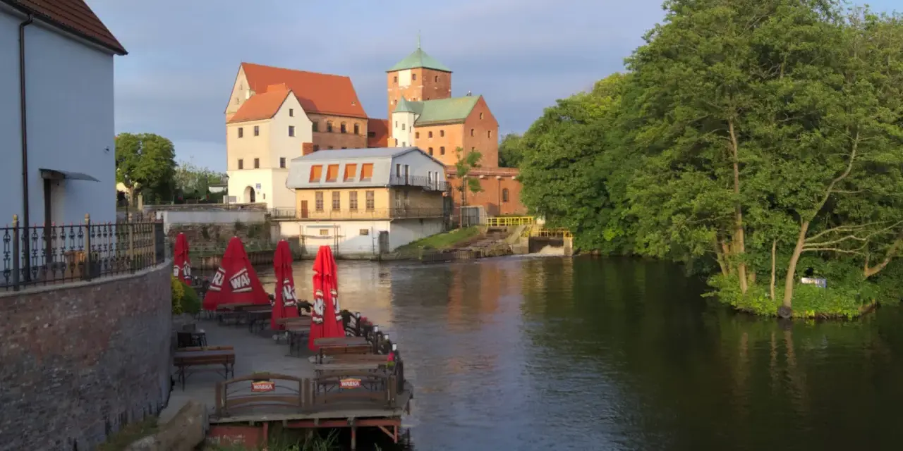 Zamek Darłowo z wieżami i zielonymi dachami, otoczony wodą i drzewami. Przy brzegu letnia restauracja z czerwonymi parasolami.