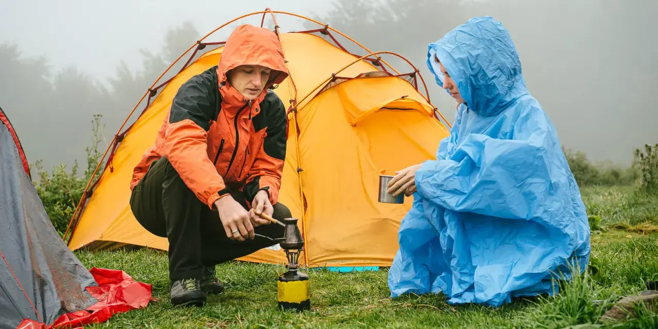 Zwei Camper in Regenponchos bereiten sich auf das Zelten bei Regen vor. Einer kocht auf einem Campingkocher, der andere hält eine Tasse.