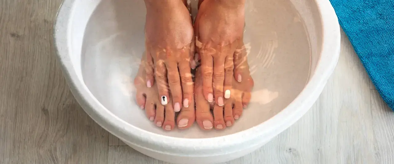 A relaxing home remedy foot soak in a white basin. Feet with french manicures and one toe nail decorated with a black and white design are submerged in clear water.