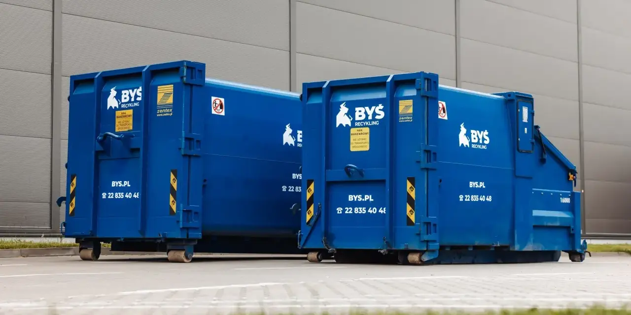 Dwa niebieskie kontenery BYS Recykling stoją przed budynkiem. Dowiedz się, ile kosztuje tona gruzu betonowego i jak go zamówić.