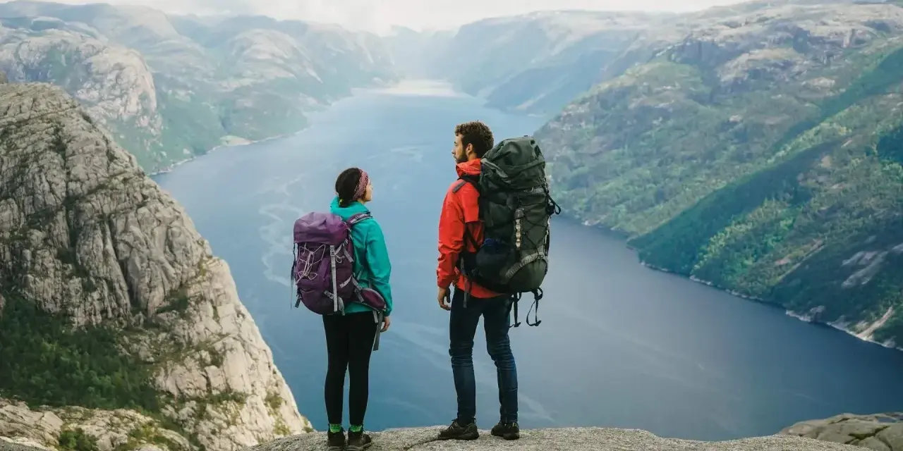Två vandrare med stora ryggsäckar står på en klippa och blickar ut över en fjord i Norge. Perfekt för camping i Norge.