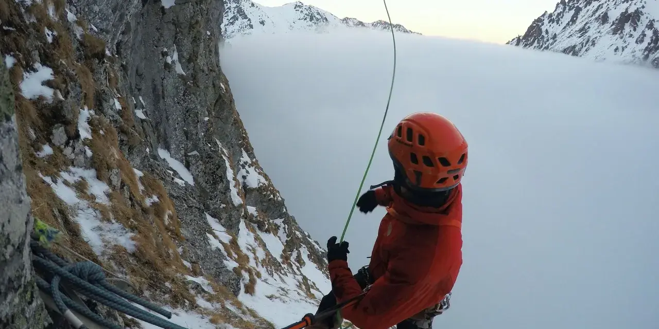 Wspinaczka górska w chmurach. Alpinista w pomarańczowym kasku i kurtce przygotowuje się do zjazdu na linie, otoczony zimowym krajobrazem i mgłą.