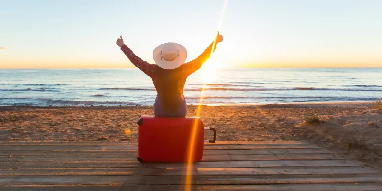Personne assise sur une valise rouge sur une plage au coucher du soleil, bras levés en signe de victoire.
