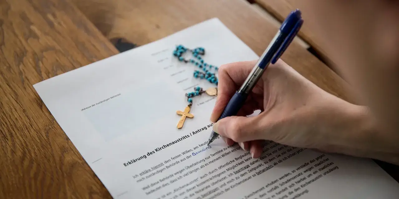 Hand unterschreibt Erklärung des Kirchenaustritts. Ab wann wirksam? Ein Rosenkranz liegt daneben.