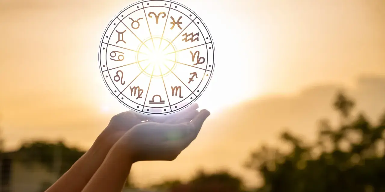 Dłonie trzymają koło zodiaku, gdzie widoczny jest symbol Wagi. Słońce przebija się przez niebo, tworząc magiczną aurę.