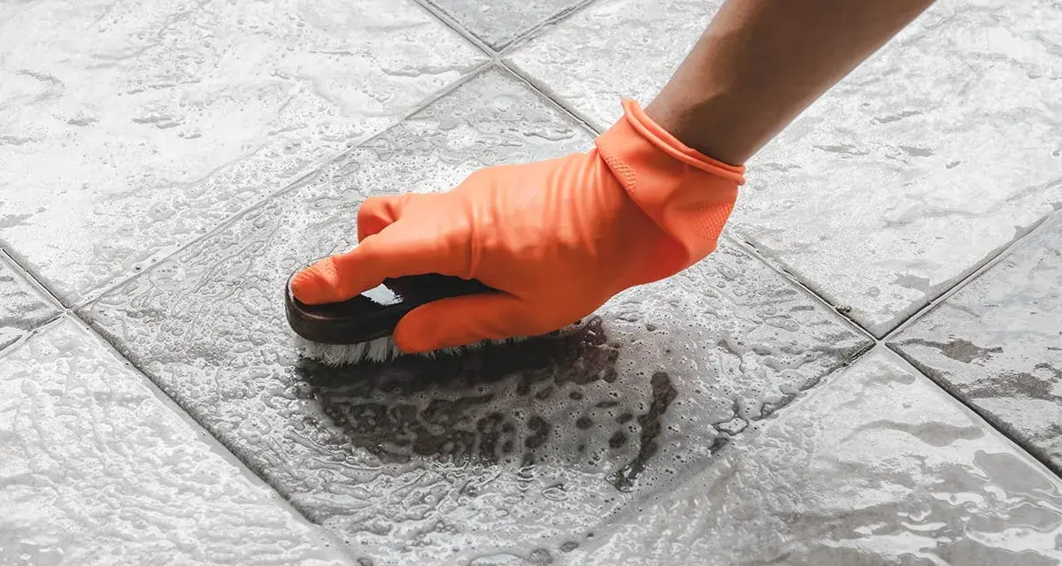 Mano con guanto arancione che spazzola un pavimento piastrellato bagnato, mostrando come togliere ruggine dal pavimento con schiuma.