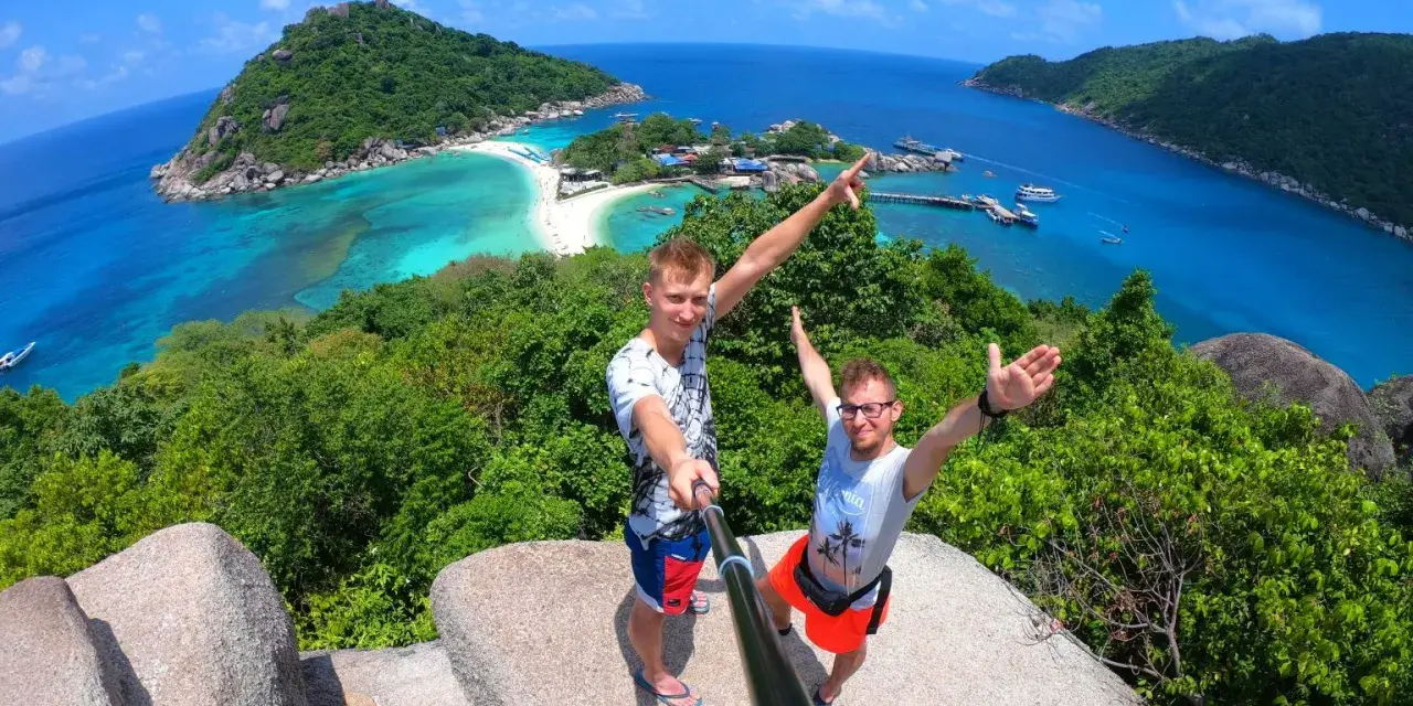 Dwóch mężczyzn robi selfie na tle tropikalnej wyspy z turkusową wodą i plażą.