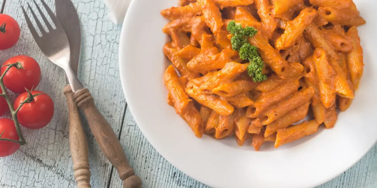 Piatto di penne alla vodka cremose, guarnite con prezzemolo, su un tavolo di legno azzurro. Accanto, pomodorini freschi e posate rustiche.
