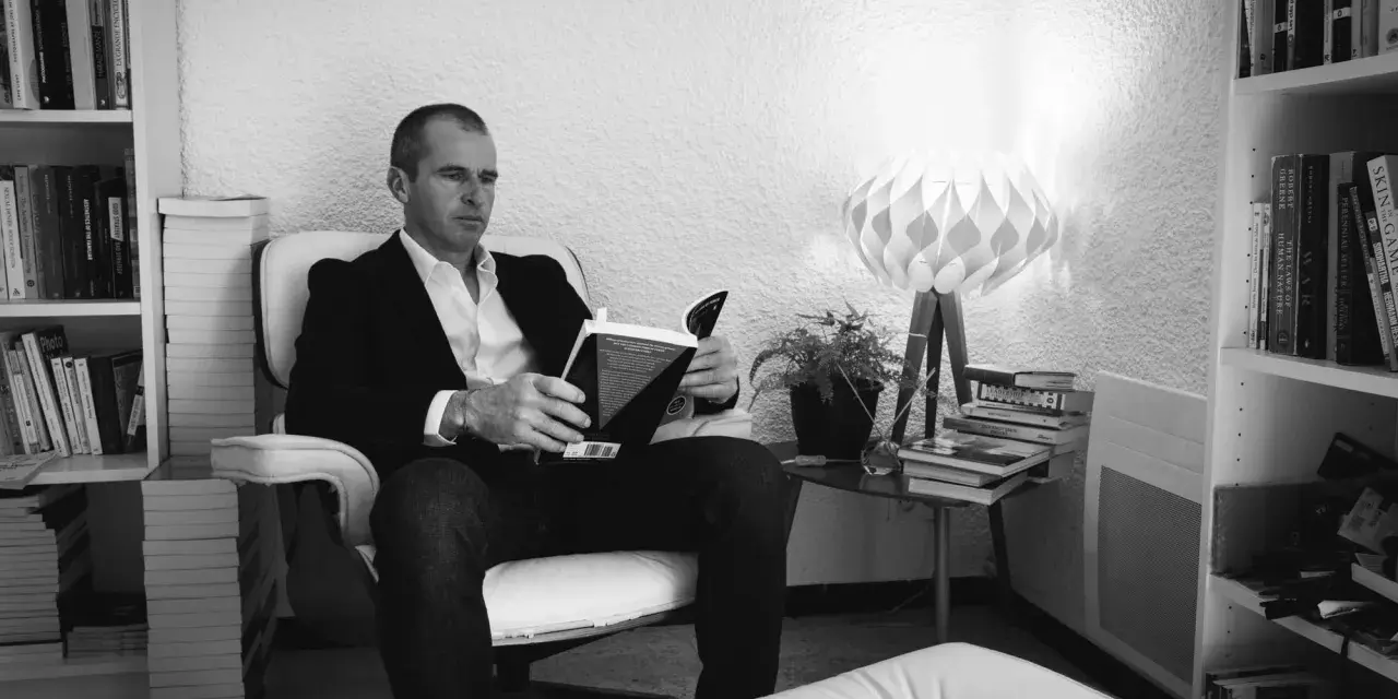 Un homme lit un livre dans un fauteuil, entouré de livres. La lumière d'une lampe éclaire la scène. ae-l af-l.