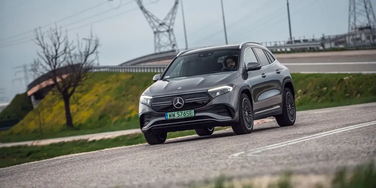 Szary Mercedes-Benz EQA jedzie po drodze, w tle linie energetyczne i drzewo.