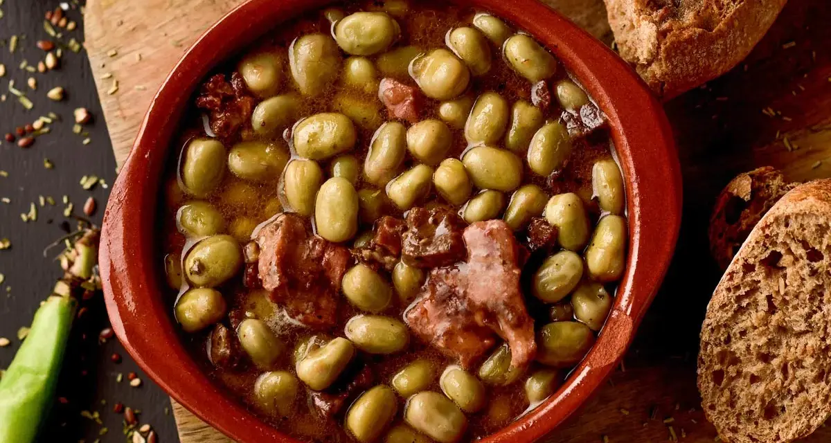 Un cuenco de barro con habas a la catalana, trozos de tocino y pan rústico al lado.