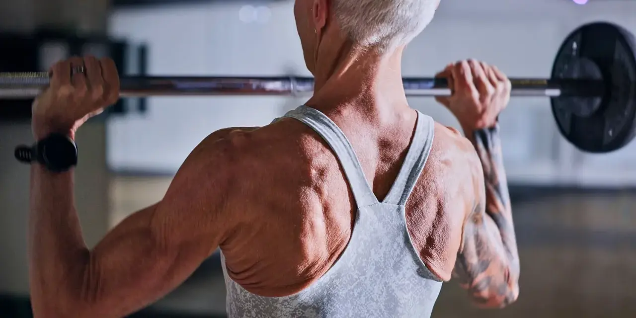 Stark ryggmuskulatur hos en kvinna som lyfter skivstång. Hon har tatuerade armar och en träningsklocka. För att bygga muskler är det viktigt med tillräckligt med gram protein per kg kroppsvikt.