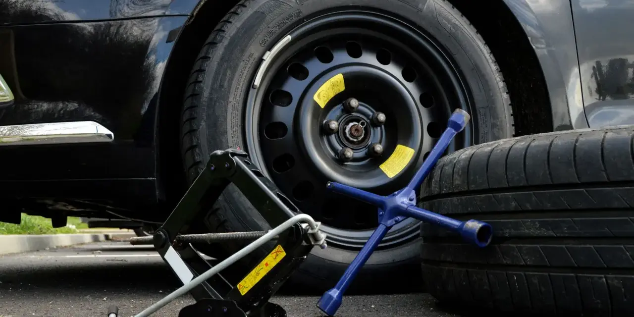 Kit per cambiare una ruota: cric, chiave a stella e ruota di scorta accanto a un'auto nera.