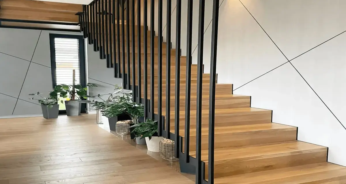 Nowoczesna klatka schodowa w domu z drewnianymi stopniami i czarną, metalową balustradą. Wokół rośliny w doniczkach.