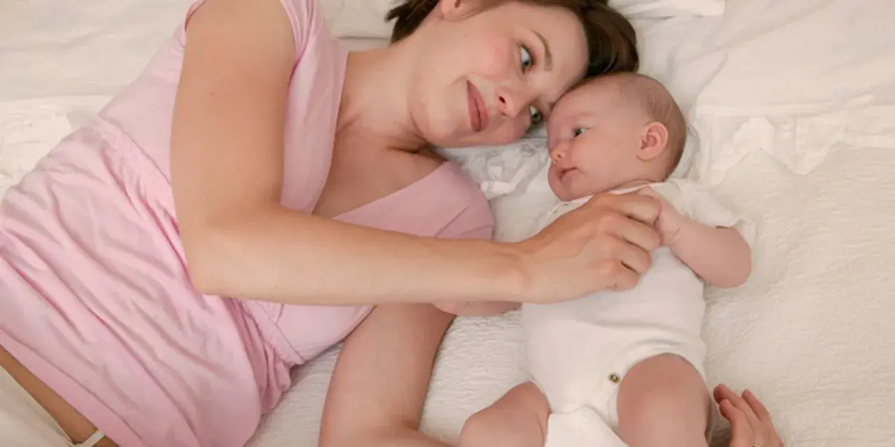 Mama patrzy z miłością na swoje 5 tygodniowe dziecko leżące obok niej na łóżku.