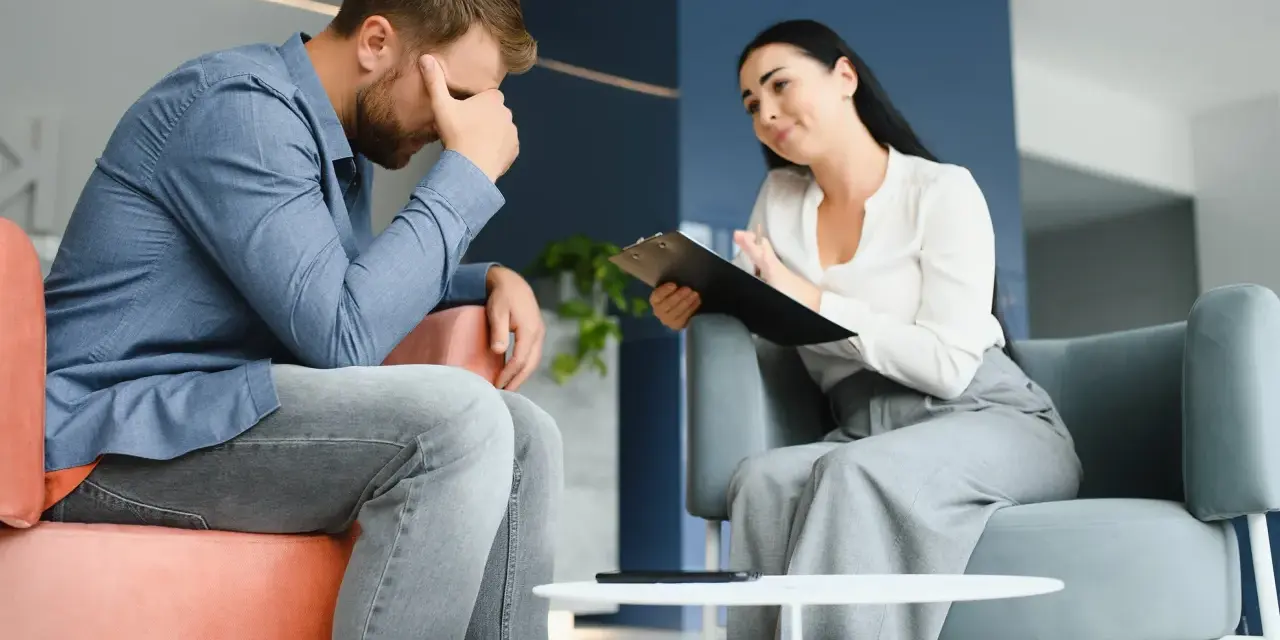 Mężczyzna w stresie podczas sesji, która może być częścią terapii uzależnień online. Terapeutaka notuje.