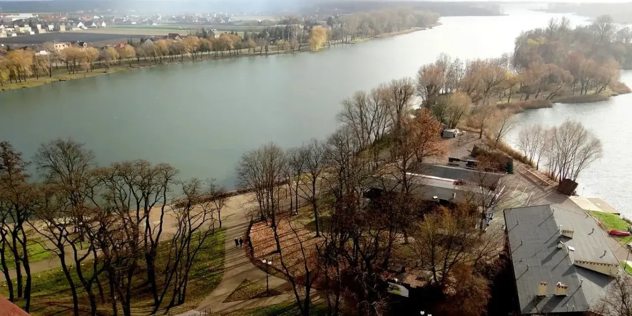 Widok na jezioro nadrowskie z wyspą i zabudowaniami. Drzewa bez liści, jesienny krajobraz.