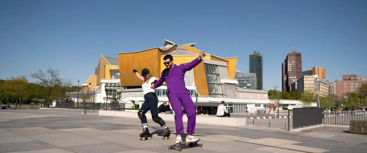Ein Mann im lila Anzug auf einem Skateboard und eine Frau auf Rollschuhen tanzen vor der Philharmonie. Ein sonniger Tag heute in Berlin.