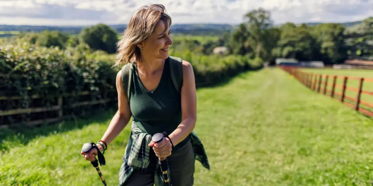 Donna sorridente con bastoncini da nordic walking, che lavora i muscoli coinvolti in un paesaggio verdeggiante.