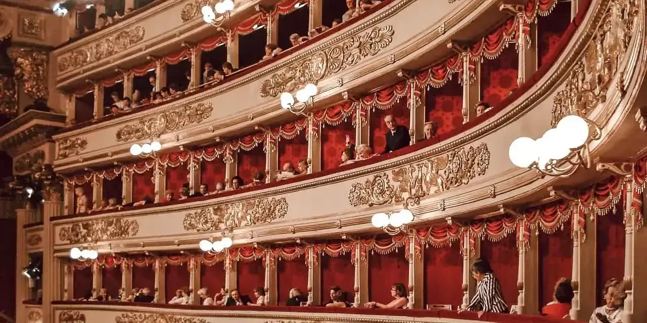 Widownia La Scali, pełna widzów w lożach, oczekuje na rozpoczęcie spektaklu. Bogaty repertuar teatru przyciąga miłośników sztuki.