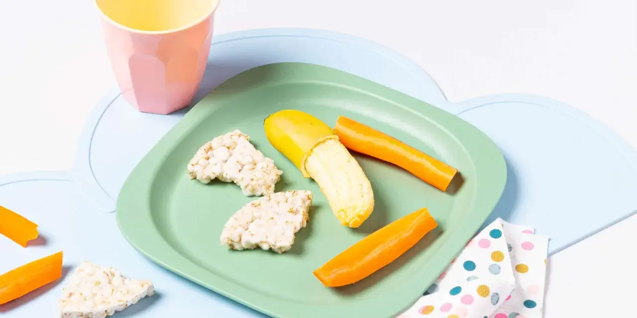 Baby-gerechte Snacks: Reiswaffeln, Karottensticks und eine halbe Banane auf einem grünen Teller.