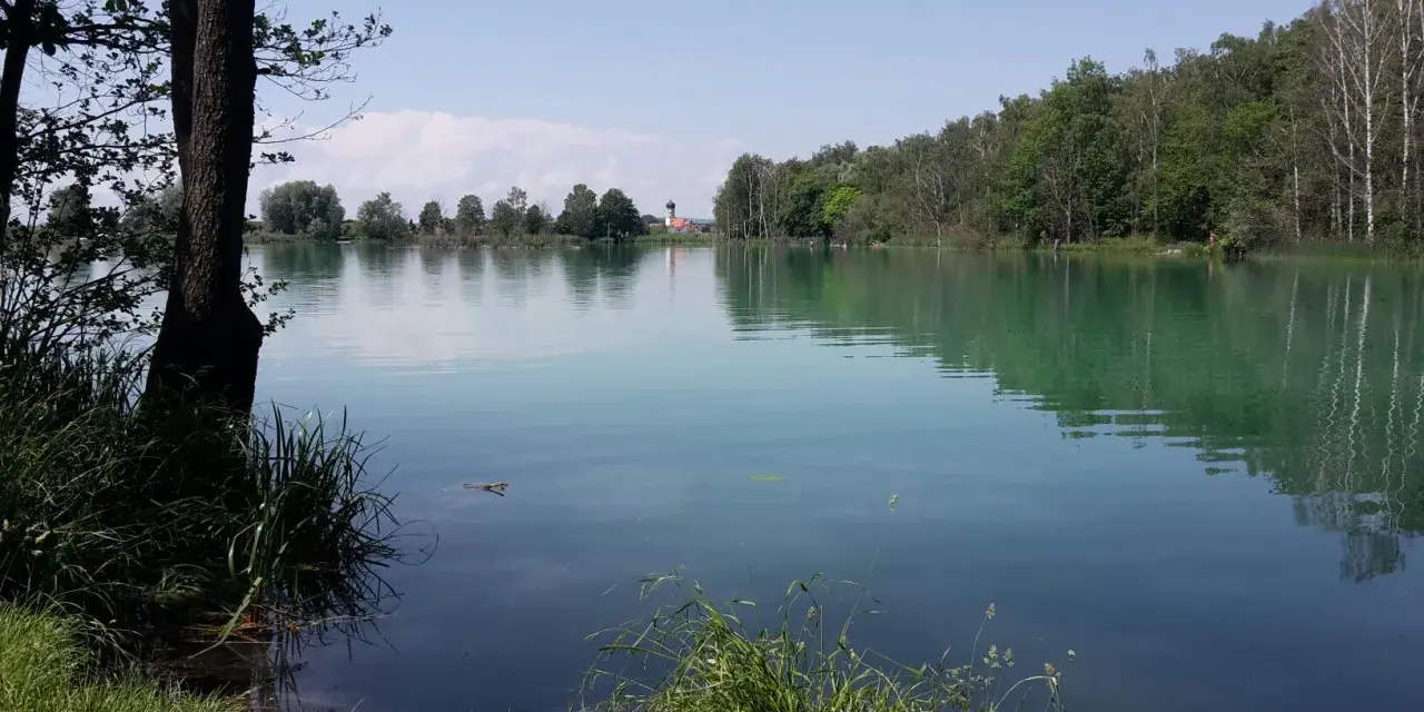 Idyllischer ersinger badesee mit grünen Ufern und einem Kirchturm in der Ferne.